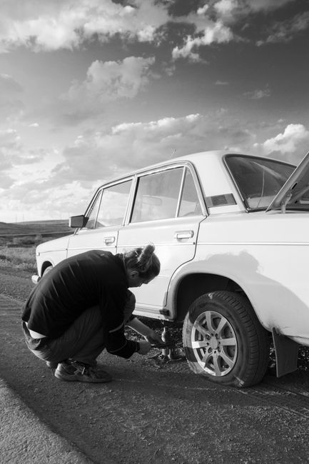 A commercial vehicle parked on the roadside with a flat tire and hazard lights blinking while traffic passes by.