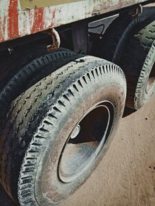 Mechanic measuring tread depth on commercial truck tires beside a parked tractor-trailer.
