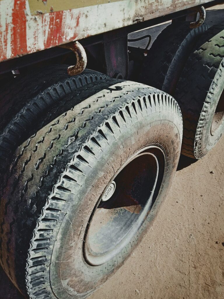 Mechanic measuring tread depth on commercial truck tires beside a parked tractor-trailer.